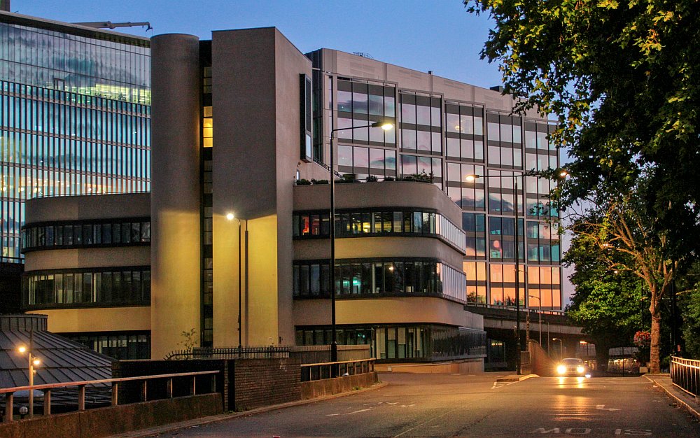 Sunset scene with the Battleship building prominent. Trees are in full leaf and a car is seen approaching the gyratory road that links Harrow Road and the Westway.