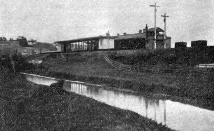 View of Higham station from the banks of the Thames and Medway canal. A signal box can be seen at the end of the station platforms and a pair of signals jut into the skyline.