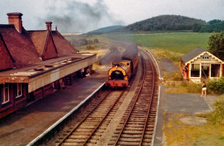 A Weybourne picture taken in 1973 around the time of the Dad's Army episode The Royal Train. The station can generally be seen and it is in a rather poor condition. The tracks curve away towards the left into the distance. There is a hill in the background which looks exactly the same even today.
