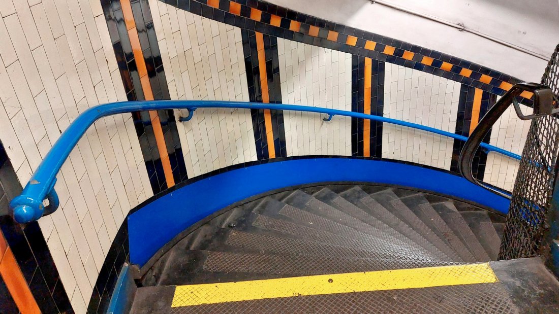 The spiral stairs at Lambeth North. The colourful tiling is obvious and its done in vertical stripes. Here it is black with amber (or gold possibly) bands. There are handrails on either side of the stairs, the left side is painted blue while that on the right seems to be a modern replacement in black.