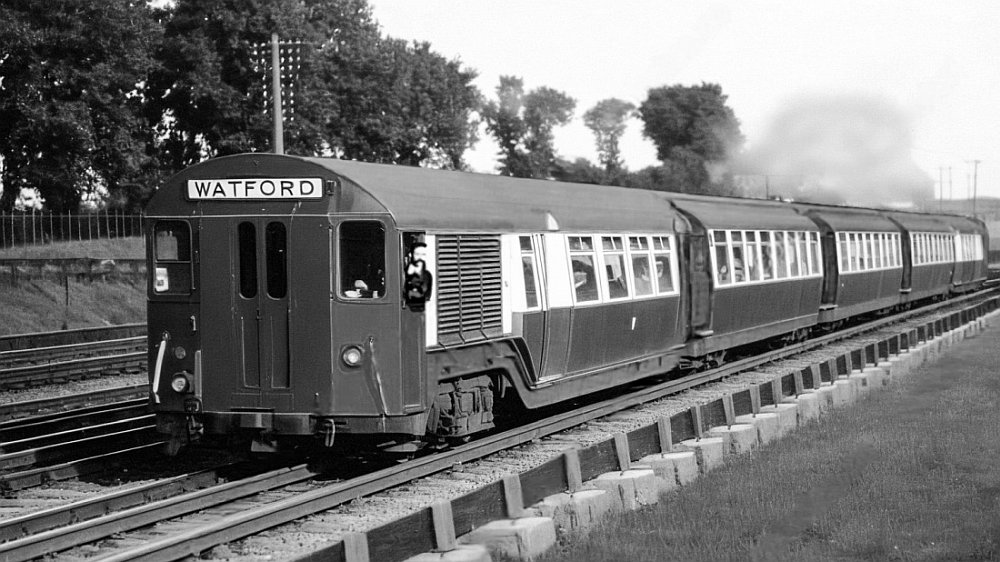 Bakerloo train heading north and seen leaving Kenton in 1917. The train's diver can be seen looking out of his window. The destination, Watford, is written in large letters on the front of the train. The main LNWR tracks are in the background behind the tube train.