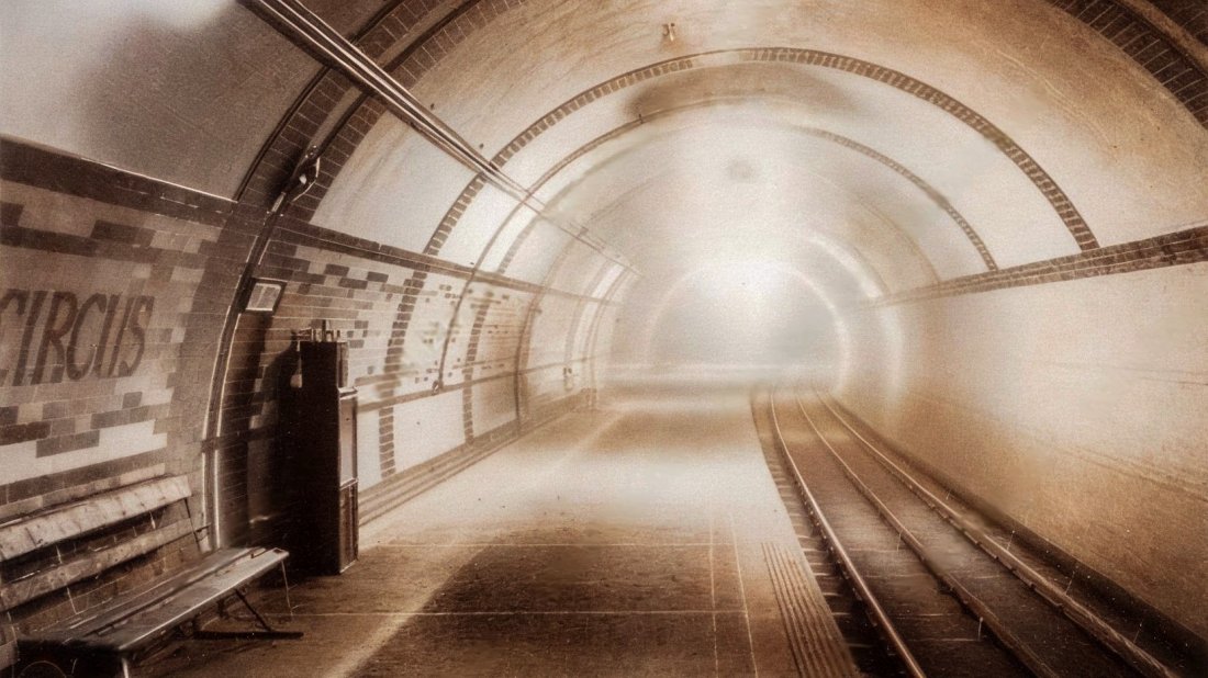 A view of the 1906 platform at Oxford Circus in a faded sepia colour. There is a seat on the left and the tracks are on the right. The station's tiled decor can be seen, with patterns along the walls and bands on the tunnel's arch.