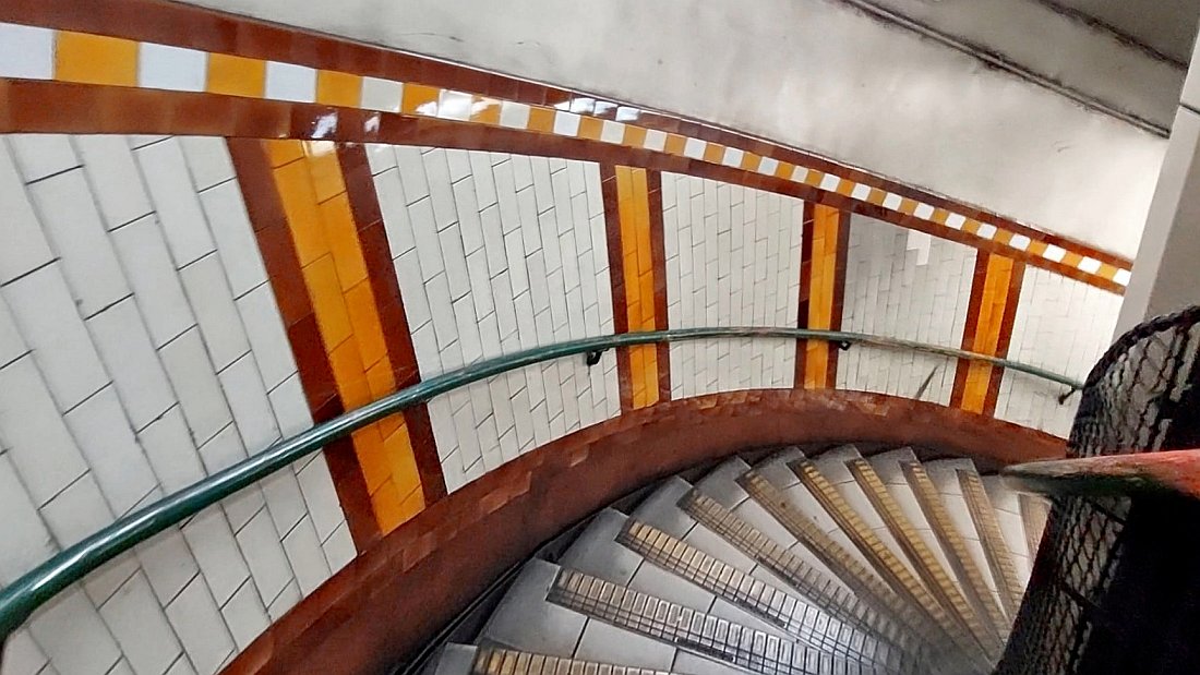 The spiral stairs at Regents Park. The colourful tiling is obvious and its done in vertical stripes. Here it is brown with amber bands. There are handrails on either side of the stairs, the left side is painted green while that on the right seems to be black with bits of faded red.
