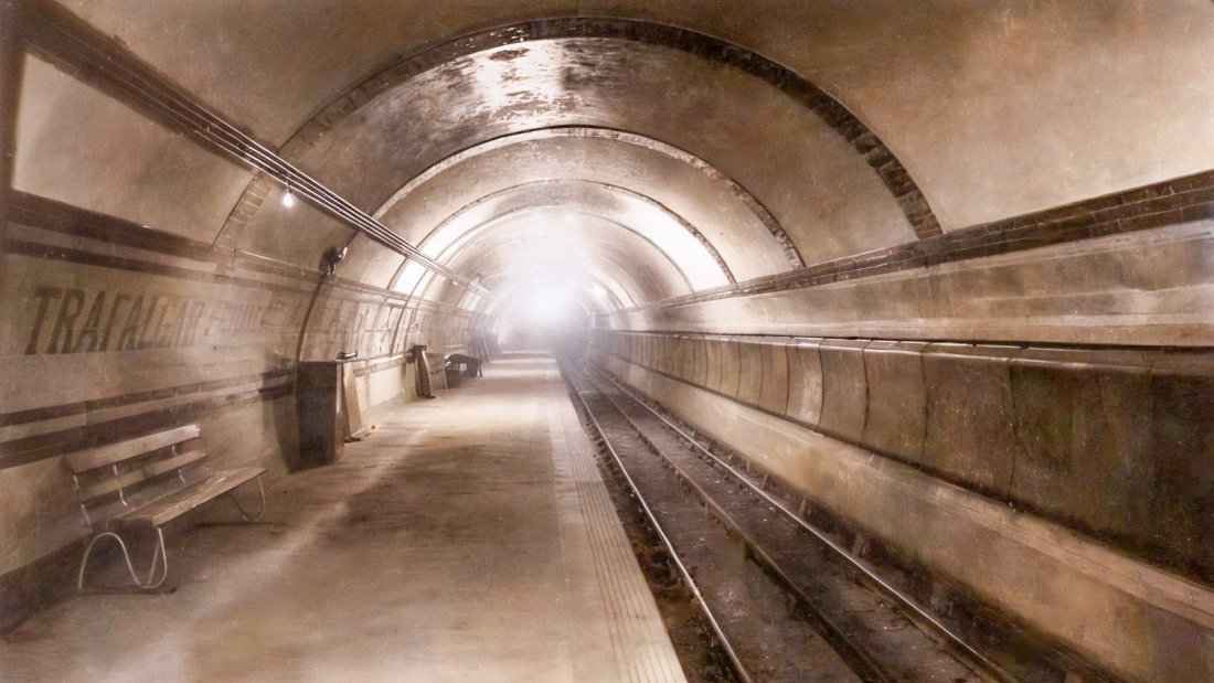A view of the 1906 platform at Trafalgar Square in a faded sepia colour. There is a seat on the left and the tracks are on the right. The station's tiled decor can be seen, with patterns along the walls and bands on the tunnel's arch. The station's name can just be seen above the seat. On the right are the cabling ducts, covered by metal sheets.